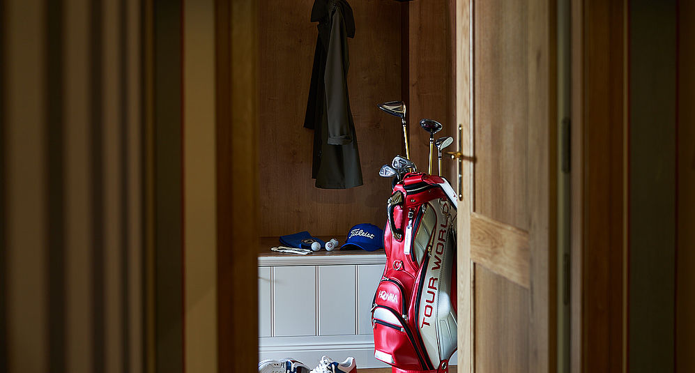 Rote Golftasche mit Schlägern steht in einer Garderobe neben Bank, Kappen und Golfbällen.