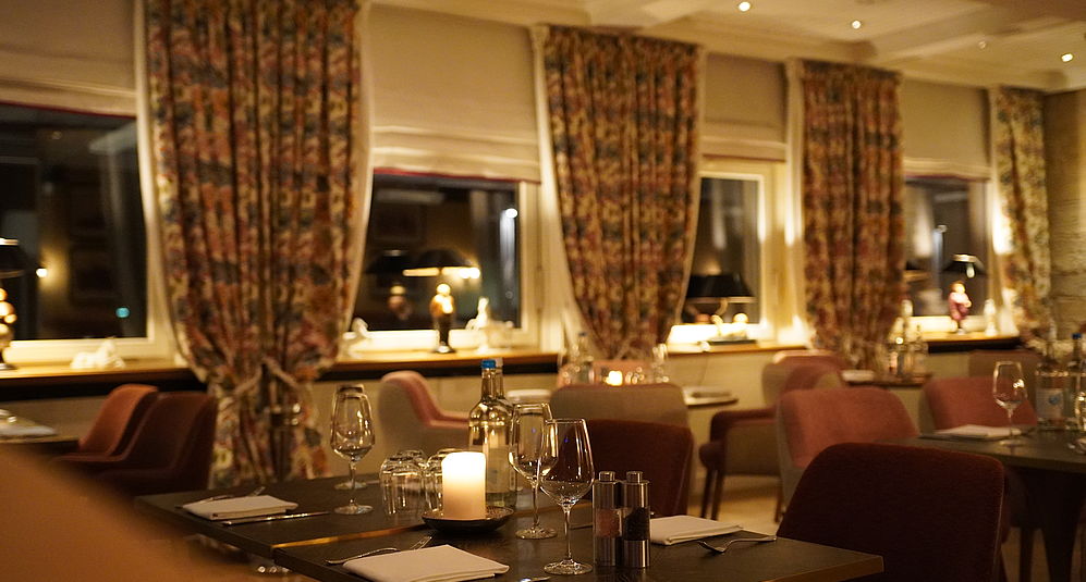 Abendlicher Restaurant-Innenraum mit gedeckten Tischen, Kerzenlicht, Gläsern und gepolsterten Stühlen.